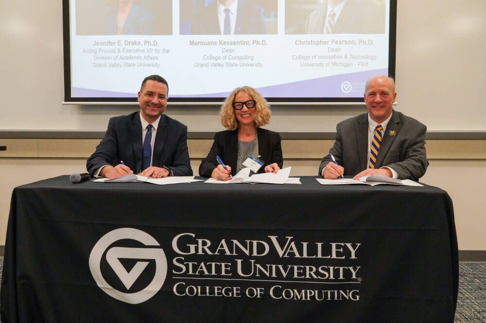 Dean Kessentini, on the left, alongside other professionals sit along a table covered with a black GVSU College of Computing cloth. They hold pens as they sign an MOU while smiling to the camera